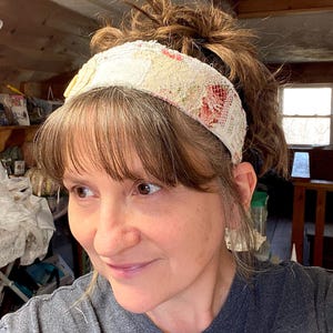 May include: A cream-colored headband with floral and lace details. The headband features a patchwork design with visible stitching and a mix of textures. The floral elements include red and pink accents, adding a pop of color to the neutral base.