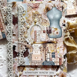 May include: A handmade journal with a floral design and lace trim. The cover features a decorative frame, a blue dress form, and a shelf with books and a wine glass. The text "adventure awaits you" is visible on the front.