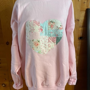 May include: A light pink long-sleeve sweatshirt featuring a large, central heart-shaped appliqué. The heart is constructed from a patchwork of various fabrics, including floral patterns, script text, and light blue and pink polka dots.