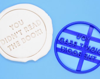May include: A blue plastic cookie cutter in the shape of a circle with the text "YOU DIDN'T READ THE BOOK!" embossed on it. The cookie cutter is on a light blue background.