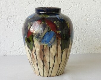 May include: A ceramic vase with a rounded body and a narrow neck. The upper portion features a dark blue glaze with red and blue floral-like patterns, while the lower section has a cream-colored base with vertical brown brush strokes.