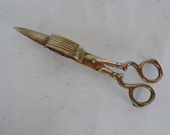 May include: Brass scissors with a decorative handle and a pointed tip. The scissors have a ribbed handle and a decorative design on the blades.