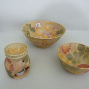 Puede incluir: Tres cuencos de cerámica con diseños florales. Los cuencos son en tonos de amarillo, naranja, rosa y verde. El cuenco más grande está en el fondo, y el cuenco más pequeño está en primer plano. Los cuencos están todos sobre una superficie blanca.