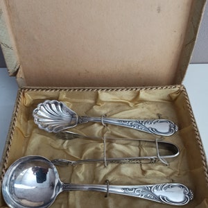 Könnte beinhalten: Ein Set aus drei silbernen Servierstücken in einer Schachtel. Die Stücke umfassen einen kleinen Löffel, eine Zange und einen größeren Löffel. Alle drei Stücke haben verzierte Designs.