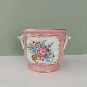 Könnte beinhalten: Ein rosa Keramik-Blumentopf mit goldenen Verzierungen und Griffen. Der Topf zeigt ein handgemaltes Blumendesign mit rosa, blauen, gelben und lila Blumen auf weißem Hintergrund. Der Topf hat einen Sockel.