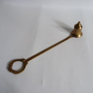 May include: A brass candle snuffer with a long handle and a bell-shaped top. The handle has a ring at the end for easy gripping.