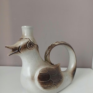 May include: A ceramic bird-shaped pitcher with a spout and handle. The pitcher is primarily off-white with brown accents on the wings and beak. The handle has a leaf-like design. The pitcher is on a white surface.
