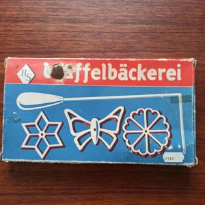 May include: Vintage waffle iron set in a box with the German text "Waffelbackerei". The box is blue and red with white outlines of a snowflake, butterfly, and flower-shaped waffle irons. The set includes a long handle.