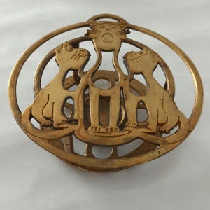 Pode incluir: Uma peça decorativa vintage em latão com tema de gato. O design apresenta três gatos estilizados dentro de uma moldura circular. Os gatos são retratados em posição sentada, com um gato central acima deles. A peça tem uma aparência envelhecida e antiga.