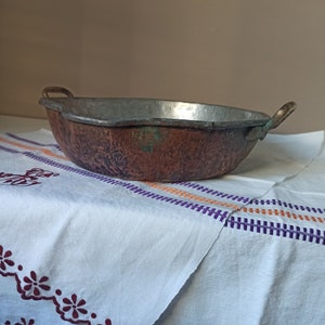 May include: A large, copper serving bowl with two handles. The bowl has a hammered texture and a worn, aged patina. It is sitting on a white tablecloth with embroidered details.
