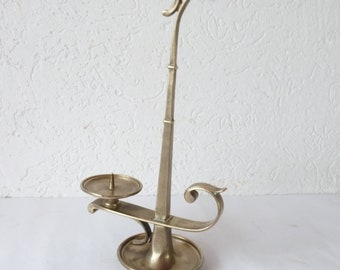 May include: A vintage brass candle holder with a unique design. It features a circular base, a tall, slender stem with a curved top, and a small dish to hold a candle. The holder has an elegant, antique aesthetic.
