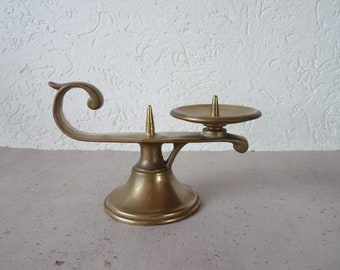 May include: A brass candle holder with a curved arm and a round base. The holder has two candle holders, one on the arm and one on the base.