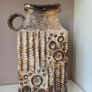 Pode incluir: Um vaso de cerâmica marrom e branco com uma superfície texturizada e um padrão geométrico. O vaso tem uma boca larga e um pescoço estreito.