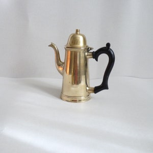 Może przedstawiać: Złoty metalowy dzbanek do kawy z czarnym plastikowym uchwytem. Dzbanek ma zaokrąglone ciało i pokrywkę z gałką na górze.