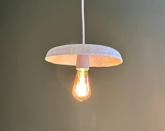 May include: A white pendant light with a round, flat shade and a single light bulb. The light is hanging from a white cord.