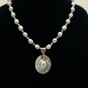 May include: A silver necklace with a large, oval-shaped pendant featuring a pearl set in a shell-like design. The necklace is made of gray pearls.