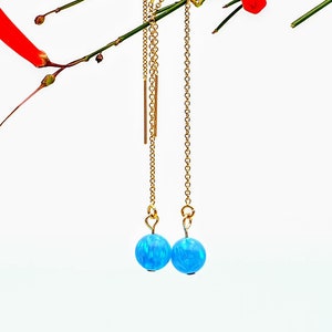 May include: A pair of gold-toned threader earrings with blue opal beads. The earrings hang from delicate chains, with a small gold bar on one earring. The earrings are displayed against a backdrop of green stems and red flowers.