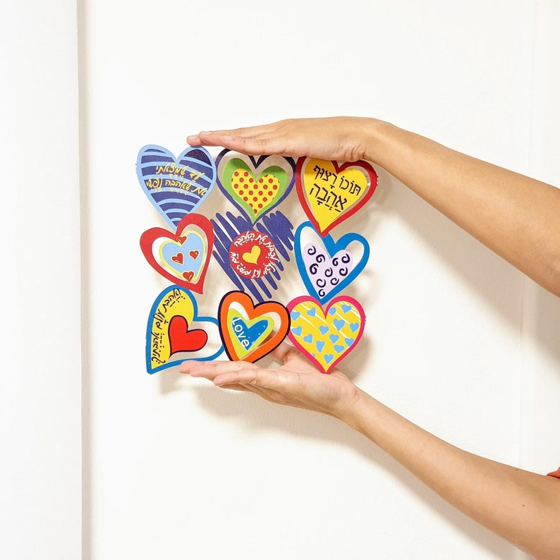 Puede incluir: Una colecci&oacute;n de corazones de papel coloridos con texto en hebreo e ingl&eacute;s, incluyendo "Love" y "Feliz cumplea&ntilde;os". Los corazones est&aacute;n dispuestos en un c&iacute;rculo y est&aacute;n sostenidos por las manos de una persona.