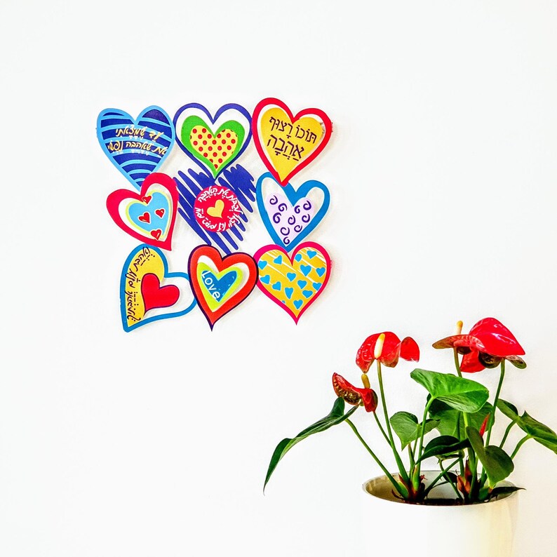 Puede incluir: Una pared blanca con nueve corazones de papel de colores pegados a ella. Los corazones tienen diferentes dise&ntilde;os y mensajes escritos en hebreo e ingl&eacute;s. Los corazones est&aacute;n dispuestos en un racimo, con el coraz&oacute;n superior siendo el m&aacute;s grande. Los corazones son todos en tonos de rojo, rosa, azul, verde y amarillo. Los corazones son una forma divertida y festiva de decorar una pared o una habitaci&oacute;n.