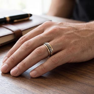 May include: A two-tone metal ring featuring a central band with engraved Hebrew text, flanked by smooth gold-colored edges. The ring has a polished finish and a wide, flat profile, worn on a finger, showcasing the detailed lettering and metallic contrast.