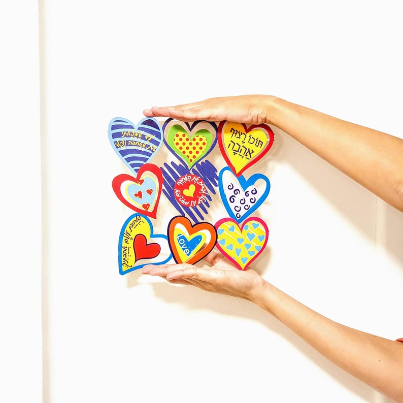Puede incluir: Una colecci&oacute;n de nueve corazones de papel coloridos con diferentes patrones y dise&ntilde;os, incluyendo corazones con texto en hebreo, un coraz&oacute;n con la palabra "Love" y un coraz&oacute;n con lunares. Los corazones est&aacute;n dispuestos en forma de cuadrado y est&aacute;n siendo sostenidos por dos manos.