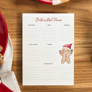 Op de afbeelding: Een kerstmaaltijdplanner-notitieblok met een peperkoekmannetje-illustratie. Het notitieblok heeft secties voor voorgerechten, hoofdgerechten, bijgerechten, desserts en een boodschappenlijst. Een rode plaat met een peperkoekmannetjeskoekje staat links.