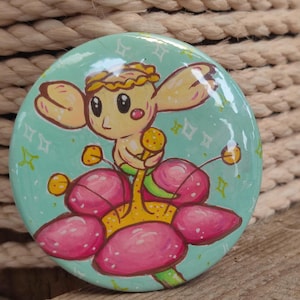 May include: A circular pin-back button featuring a glossy finish and a light blue background. The illustration depicts a small, pale, fairy-like creature with large eyes and a flower-like head, sitting atop a large, vibrant pink flower blossom.