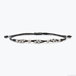 Op de afbeelding: Een zwarte armband van touw met zilveren kralen. De kralen zijn klein en rond en zijn gerangschikt in een clusterpatroon.