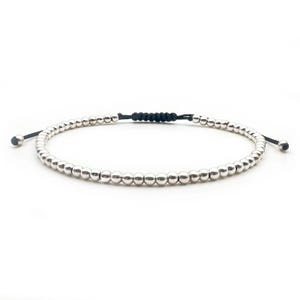 May include: Silver beaded bracelet with a black adjustable cord. The bracelet features small, round silver beads strung together, with a black woven section in the centre. The ends have small silver beads and black cords for adjusting the size.