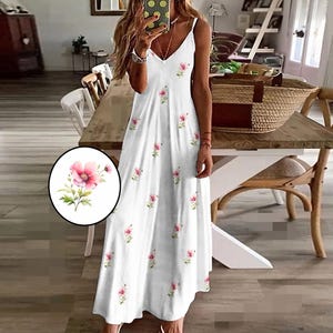 Op de afbeelding: Witte maxi-jurk met een bloemenpatroon. De jurk heeft een V-hals en spaghettibandjes. Roze bloemen zijn over de stof verspreid. De jurk is vloerlang en lijkt van een licht materiaal te zijn.