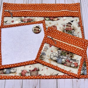 Peut inclure: Trois pochettes zippées en tissu orange et blanc avec des fenêtres en vinyle transparent. Les pochettes présentent un imprimé sur le thème de l'automne avec des citrouilles, des courges et un épouvantail. La plus grande pochette a un insert en feutre blanc.