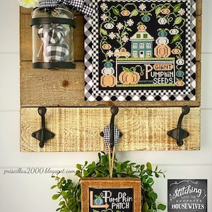 Peut inclure: Une étagère murale en bois avec un motif de point de croix encadré noir et blanc représentant une maison, des citrouilles et le texte "Giant Pumpkin Seeds". Un bocal en verre avec un tissu à carreaux noir et blanc à l'intérieur est suspendu à l'étagère. Une petite pancarte en bois avec un motif de point de croix noir et blanc représentant des citrouilles et le texte "Pumpkin Patch" est suspendue à un crochet sur l'étagère. Une petite pancarte en ardoise noire avec le texte "Stitching with the Housewives" est sur l'étagère.