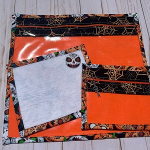 Peut inclure: Trois pochettes orange à fermeture éclair avec un motif d'araignée noir et orange. La plus grande pochette a une fenêtre en plastique transparent. La pochette du milieu a une doublure en tissu blanc. La plus petite pochette a une fermeture éclair sur le dessus.