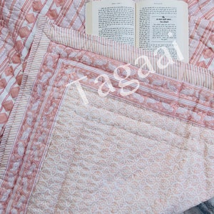 May include: A pink and white cotton quilt with a paisley border and a geometric pattern. The quilt is folded in half and a book is lying on top of it.