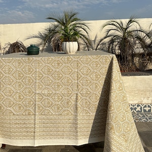 Pode incluir: Uma toalha de mesa retangular com um padrão floral dourado e branco repetido. A toalha de mesa está sobre uma mesa, com um pote de cerâmica verde e um vaso de cerâmica branco em cima. Palmeiras e céu azul ao fundo.