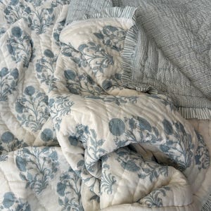 Op de afbeelding: Een gewatteerde sprei met een bloemenpatroon in blauw- en wittinten. De sprei heeft een herhalend ontwerp van gestileerde bloemen en bladeren. De randen zijn afgewerkt met een gestreepte stof en een gefranjerde rand. Het beddengoed is zacht en uitnodigend.