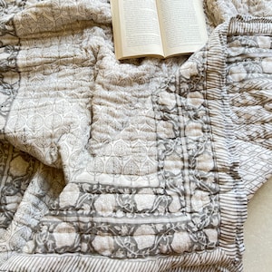 Pode incluir: Um edredom estampado em branco e cinza com um livro em cima. O edredom tem um design floral e uma borda de listras.