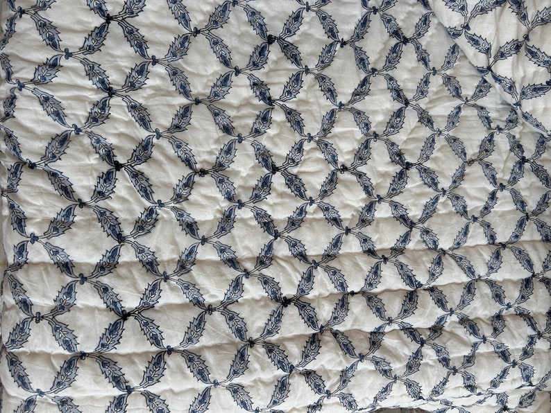 K&ouml;nnte beinhalten: Wei&szlig;er Stoff mit einem sich wiederholenden Muster aus blauen, stilisierten Bl&auml;ttern in einer Diamantform.