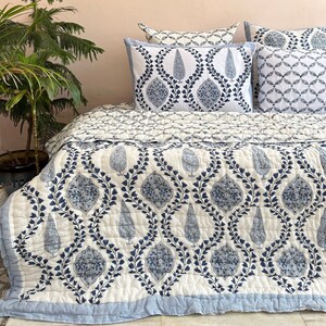 K&ouml;nnte beinhalten: Eine wei&szlig;e und blaue Steppdecke mit floralem Muster. Die Steppdecke ist aus Baumwolle und hat einen hellblauen Rand. Die Steppdecke ist gefaltet und auf einem Bett mit passenden Kissenbez&uuml;gen ausgestellt.