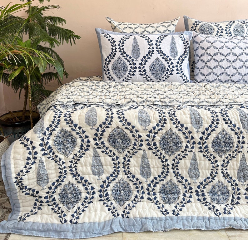 K&ouml;nnte beinhalten: Eine wei&szlig;e und blaue Steppdecke mit floralem Muster. Die Steppdecke ist aus Baumwolle und hat einen hellblauen Rand. Die Steppdecke ist gefaltet und auf einem Bett mit passenden Kissenbez&uuml;gen ausgestellt.