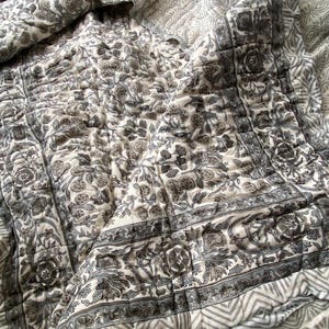 May include: A grey and white floral patterned quilt. The quilt has a border with a geometric design. The design is soft and inviting, suitable for a bedroom or living space. The quilt is made of a soft material.