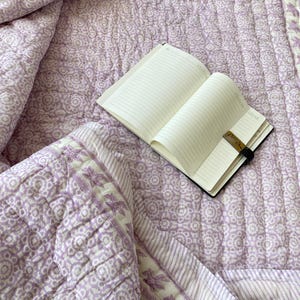 Könnte beinhalten: Eine hellviolette und weiße Steppdecke mit einem sich wiederholenden Blumen- und Geometriemuster, einem passenden gefalteten Teil und einem offenen, linierten Notizbuch darauf. Das Notizbuch hat einen schwarzen Gummibandverschluss mit einer kleinen braunen Lasche.