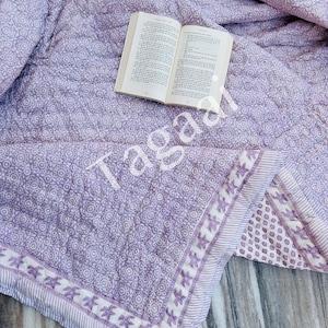 Op de afbeelding: Een lavendelkleurige en witte katoenen deken met een bloemenprint en een witte rand. De deken is gevouwen op een houten oppervlak met een boek erop.