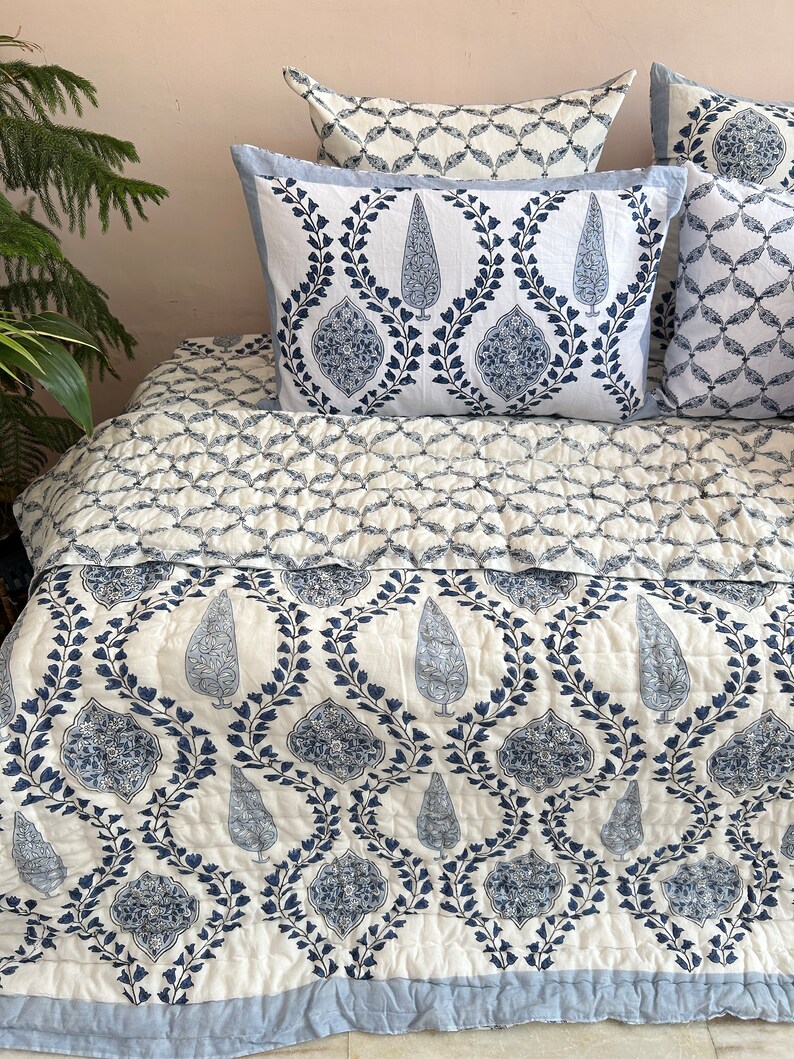 K&ouml;nnte beinhalten: Eine wei&szlig;e Baumwolldecke mit einem blauen Blumenmuster. Die Decke ist gefaltet und auf einem Bett mit passenden Kissenbez&uuml;gen ausgestellt.