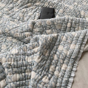 May include: A quilted blanket featuring a light blue and white floral block print pattern. The fabric has a textured, puckered surface and is finished with a ruffled edge featuring thin vertical stripes. A small black rectangular book rests on top.