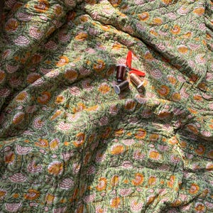 May include: A green floral quilt with a repeating pattern of orange and pink flowers. The quilt has a quilted texture and is folded. Three spools of thread in brown and gold are on the quilt.