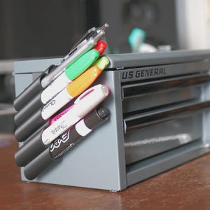 Op de afbeelding: Een grijze metalen gereedschapskist met de tekst "US GENERAL" aan de zijkant, met een verzameling Sharpie-markers en een pen. De markers zijn in verschillende kleuren, waaronder rood, groen, oranje, roze en zwart. De markers zijn gerangschikt in een houder die aan de zijkant van de kist is bevestigd.