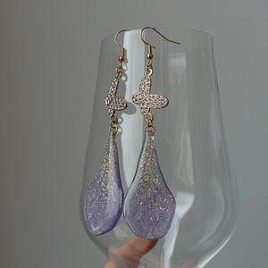 Può includere: Orecchini pendenti con motivo a farfalla. Gli orecchini presentano ganci color oro e decorazioni a farfalla con strass. I pendenti a goccia sono color lavanda con glitter, per un look delicato ed elegante.