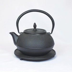 Op de afbeelding: Een zwarte gietijzeren theepot met een getextureerd oppervlak en een klein knopje op de deksel. De theepot staat op een zwarte metalen standaard.
