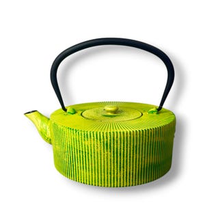 Può includere: Una teiera rotonda in ghisa con una finitura testurizzata verde lime e gialla brillante con scanalature verticali. La teiera include un coperchio circolare abbinato con un piccolo pomello e un robusto manico ad arco in metallo nero.
